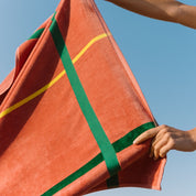 Cognac beach towel with green and yellow stripes held by a person against a clear blue sky.