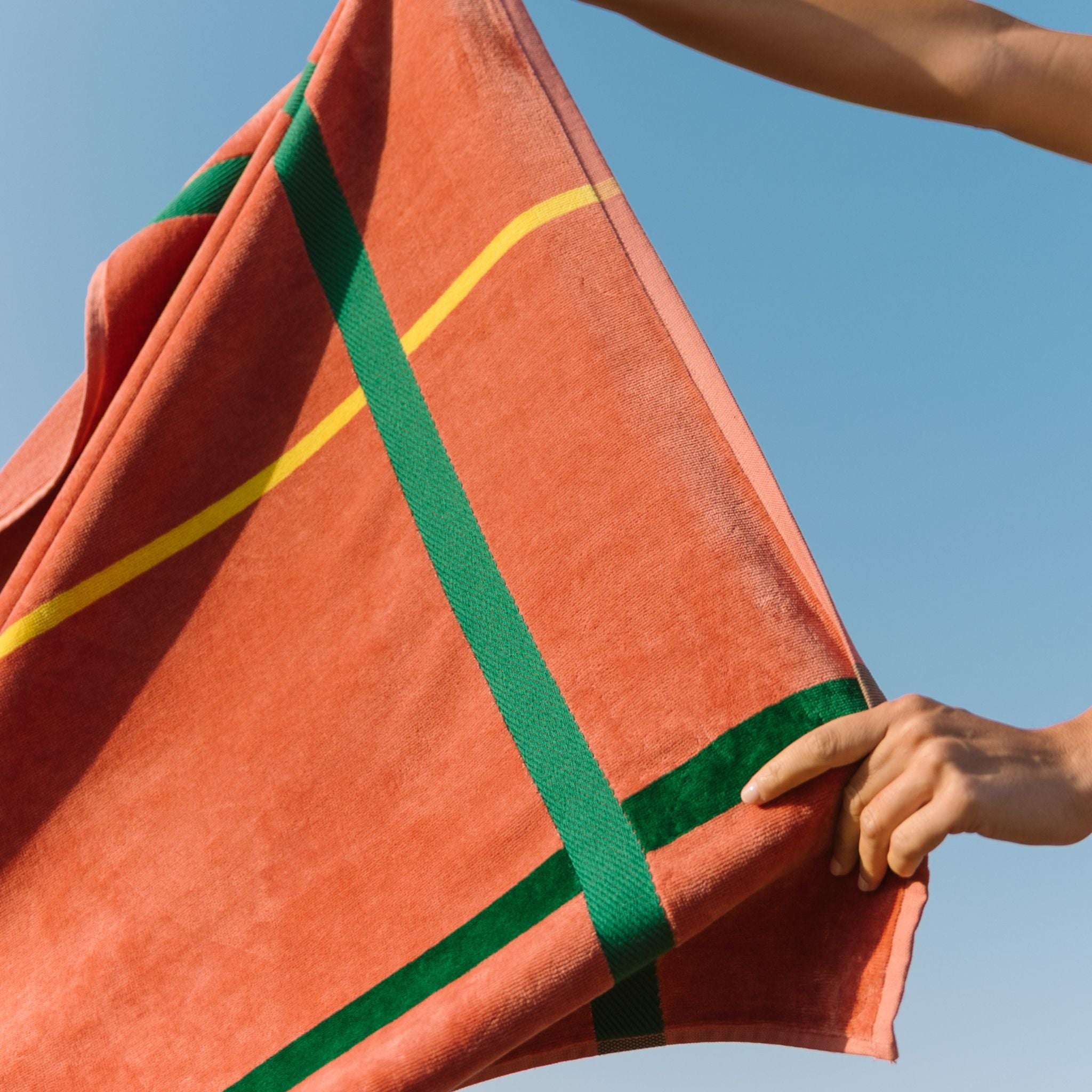 Cognac beach towel with green and yellow stripes held by a person against a clear blue sky.