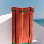 Cognac beach towel with green and yellow stripes on a white surface with blue sky and ocean in the background