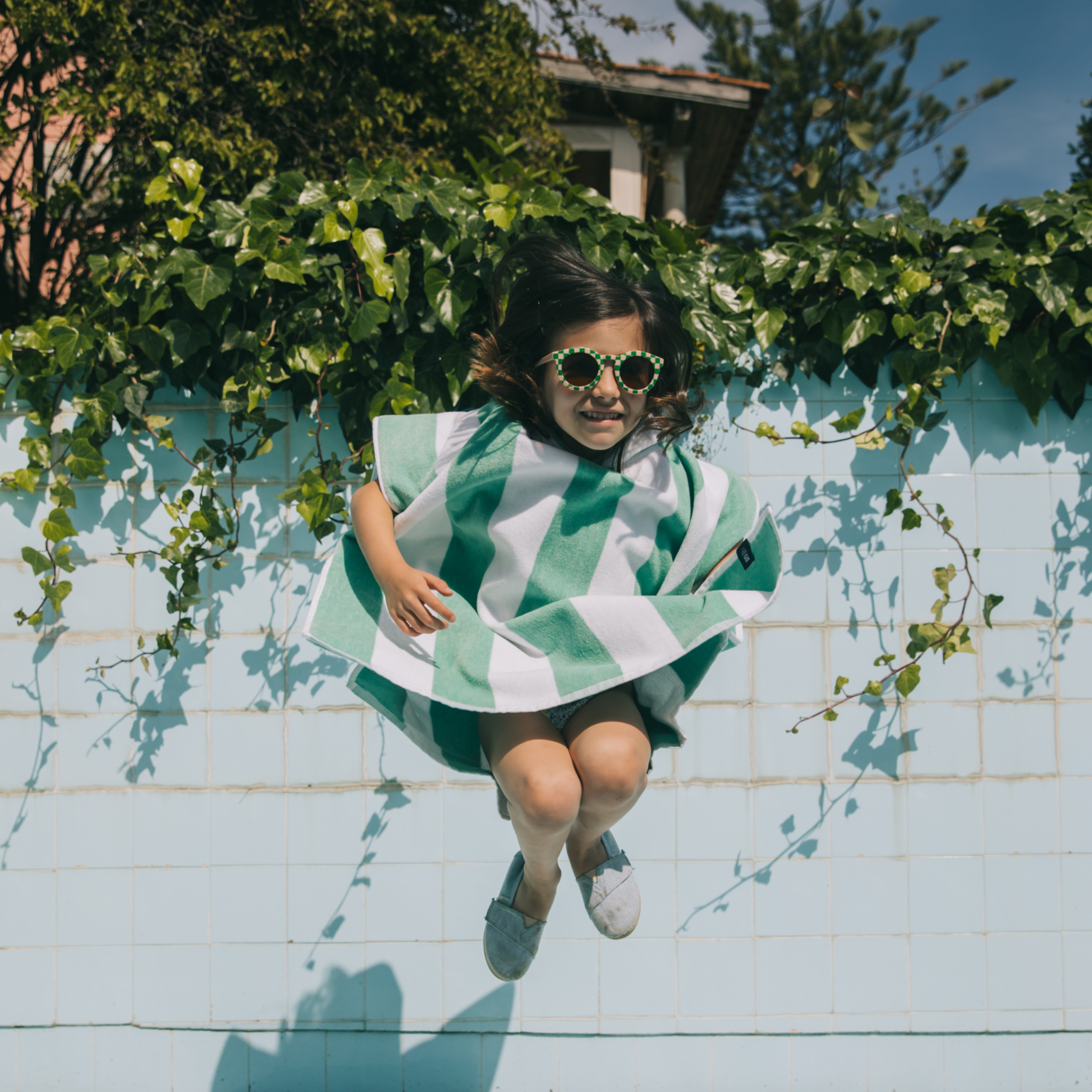Child wearing a green and white striped poncho and sunglasses, standing on a tiled pool deck with plants around.