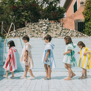 A group of children standing by a poolside wearing colorful ponchos in blue, green, pink, yellow, beige, and gray.