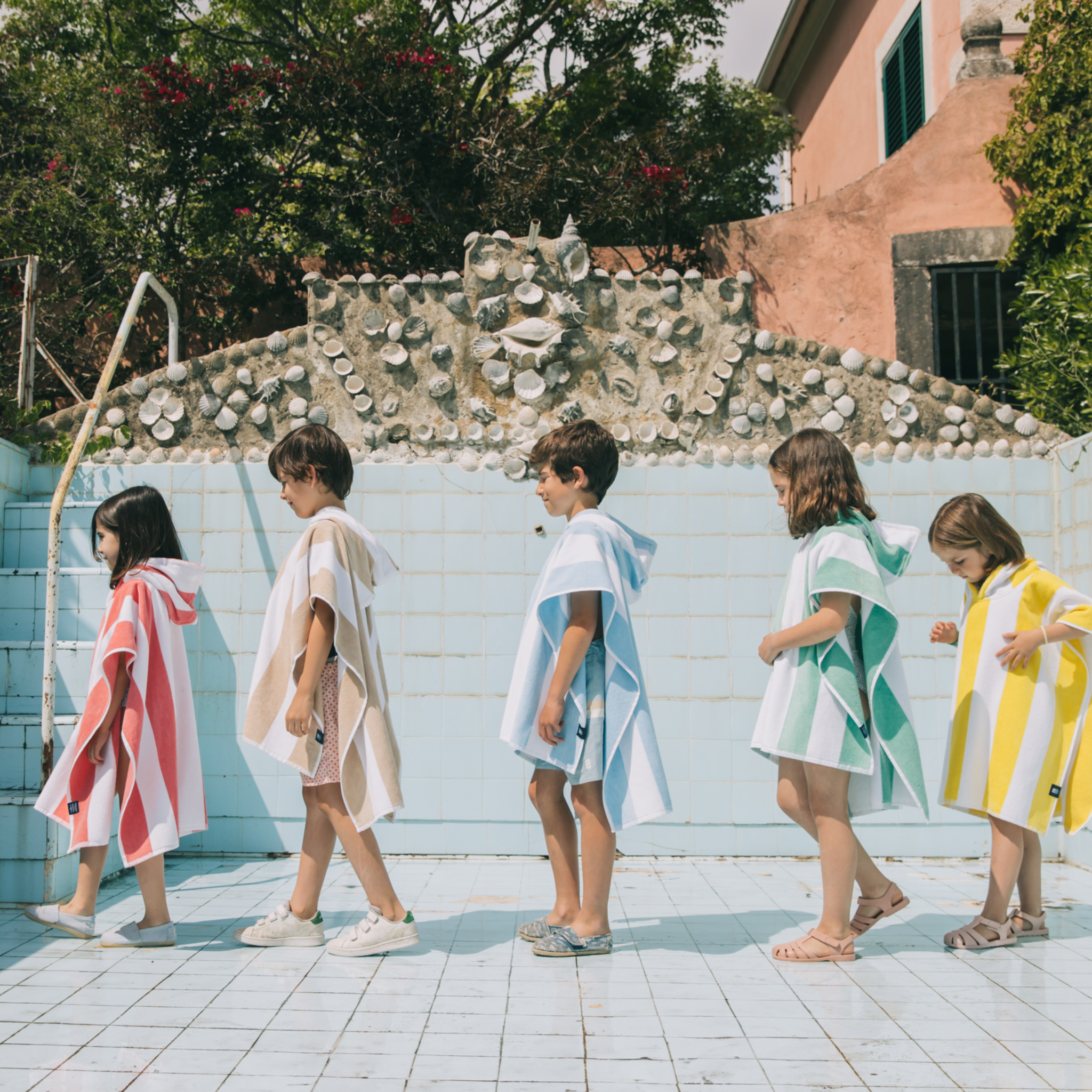 A group of children standing by a poolside wearing colorful ponchos in blue, green, pink, yellow, beige, and gray.