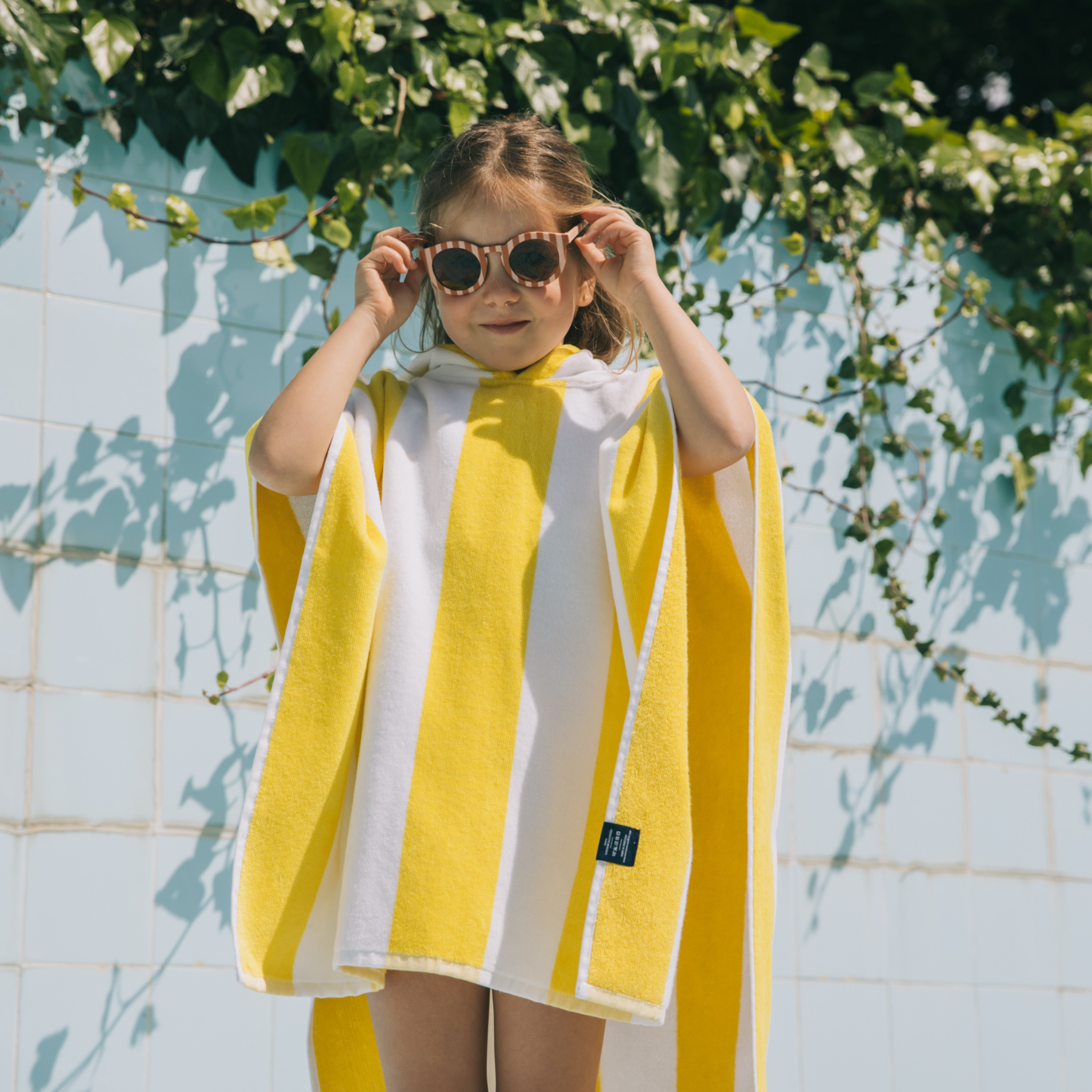 Child wearing a yellow and white poncho with sunglasses, standing against a tiled wall with greenery.