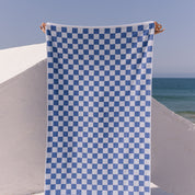 Blue checkered beach towel held up against a clear blue sky and ocean.