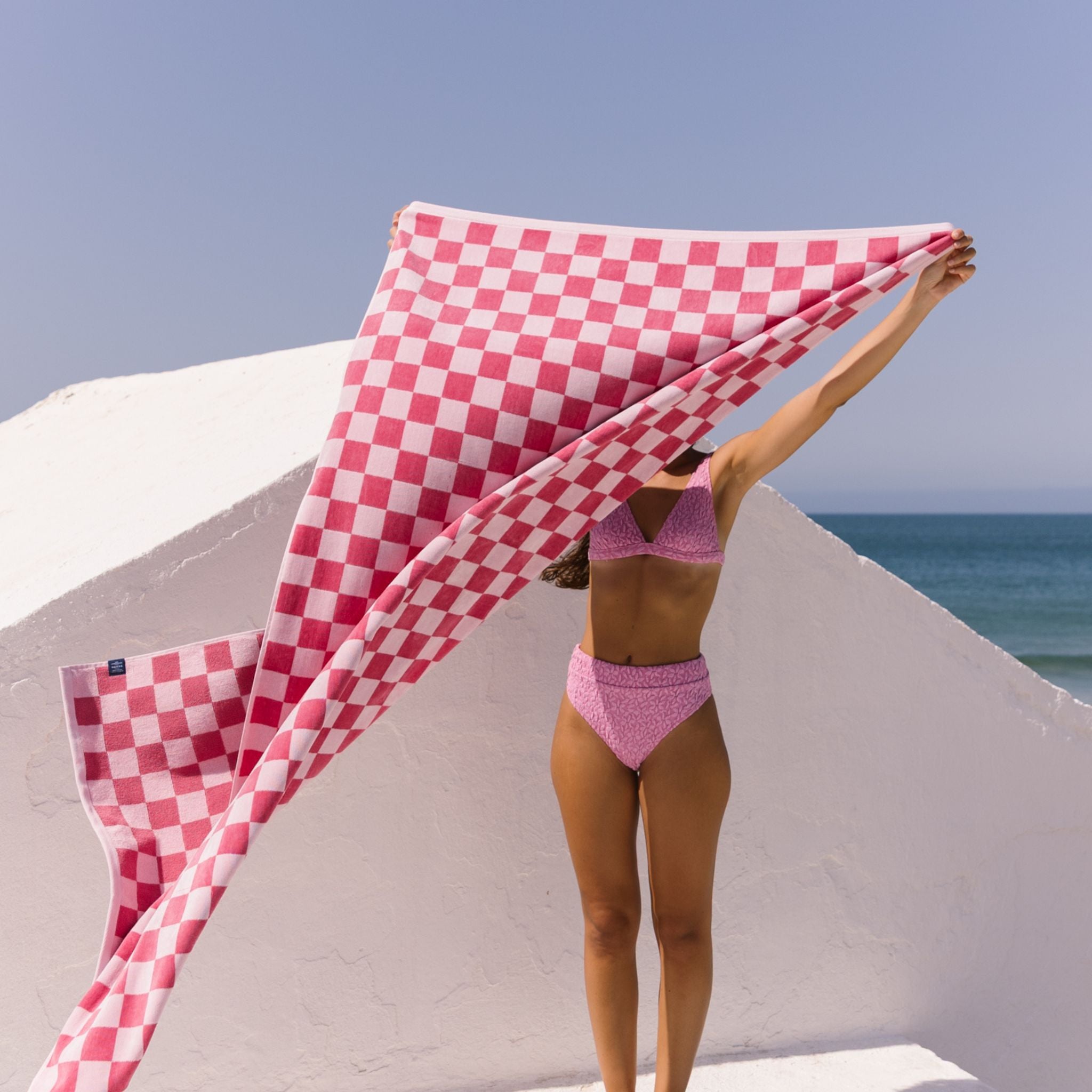 Woman in pink bikini holding a pink checkered towel against a beach backdrop