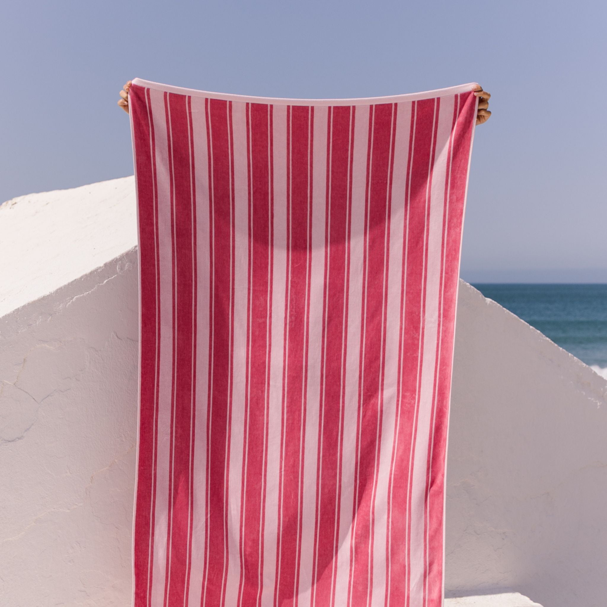 Pink striped beach towel held up against a clear blue sky and ocean.