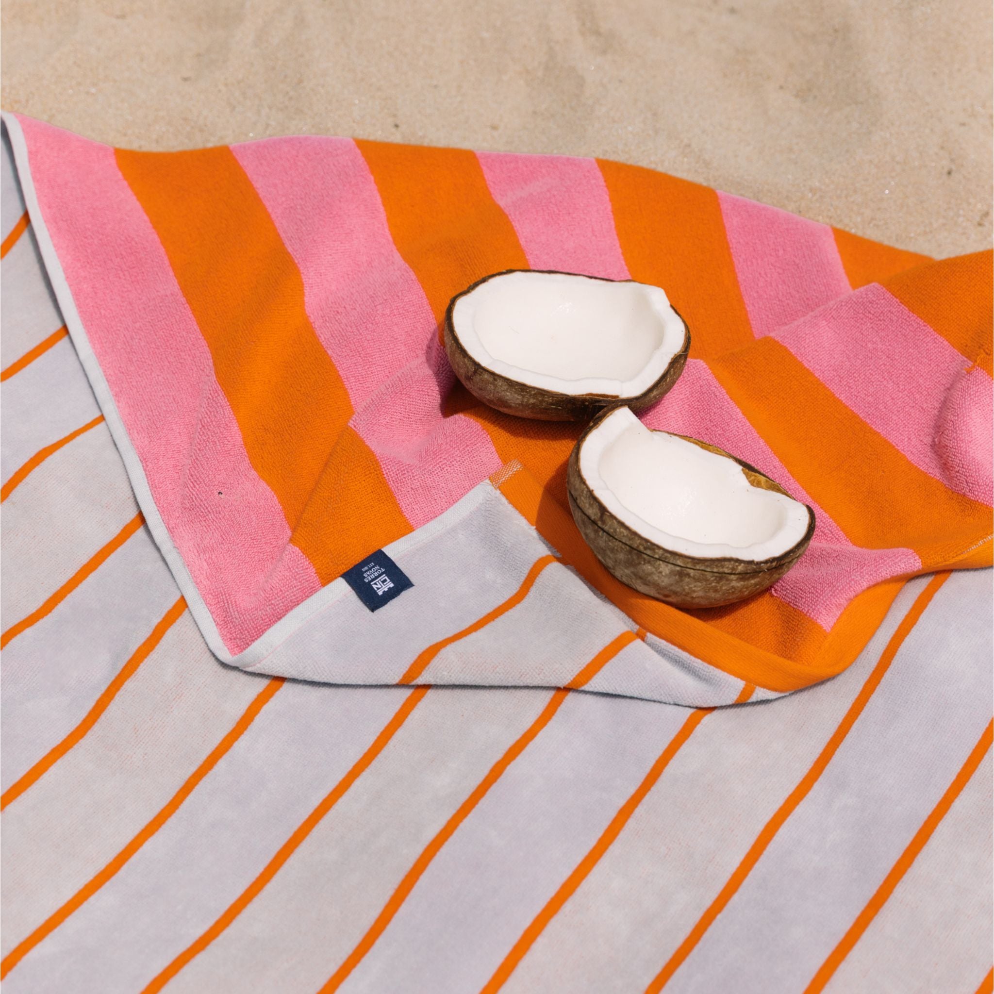 A beach towel with vertical stripes in orange, pink, and gray, laid out on the sand with a coconut cut in half on top.