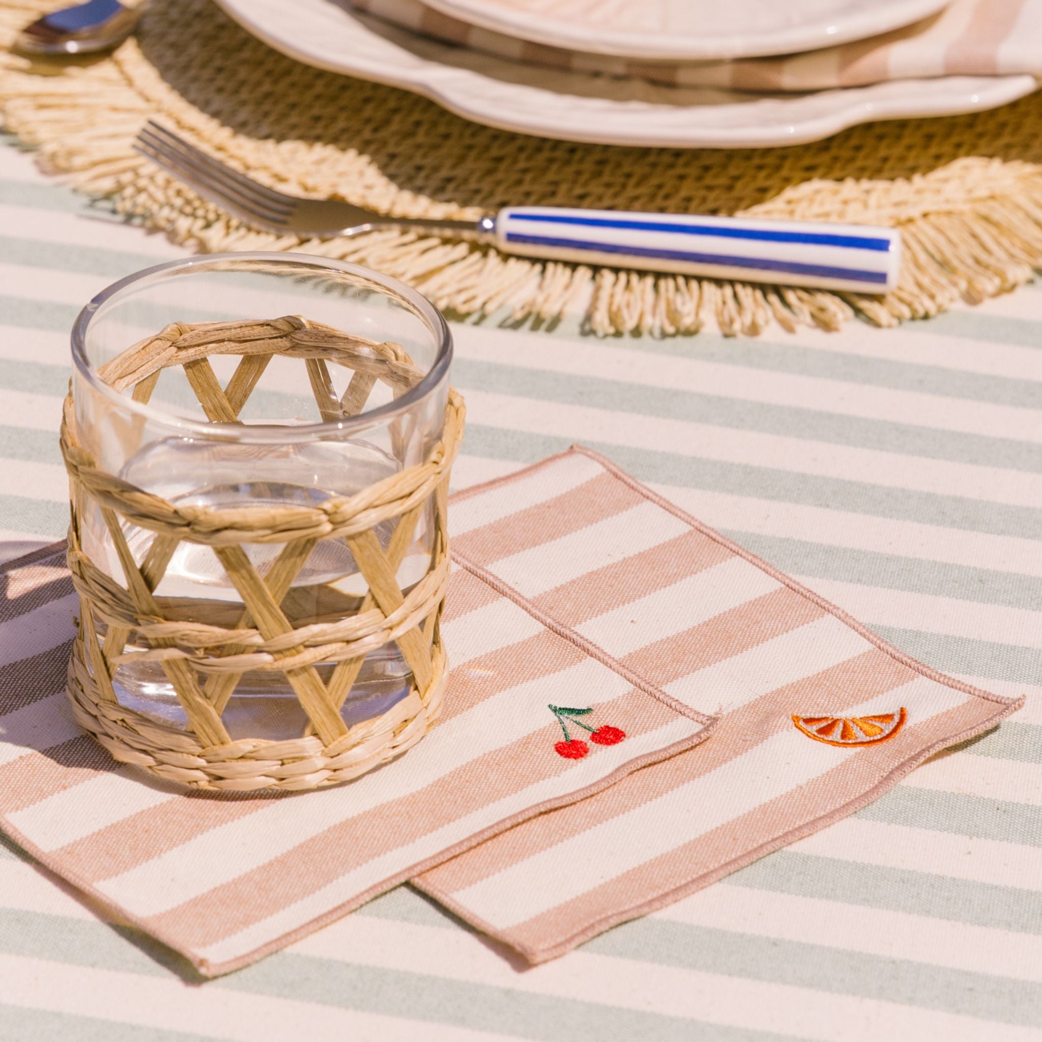 Woven glass on a striped tablecloth with a striped napkin featuring embroidered designs of orange and cherry.