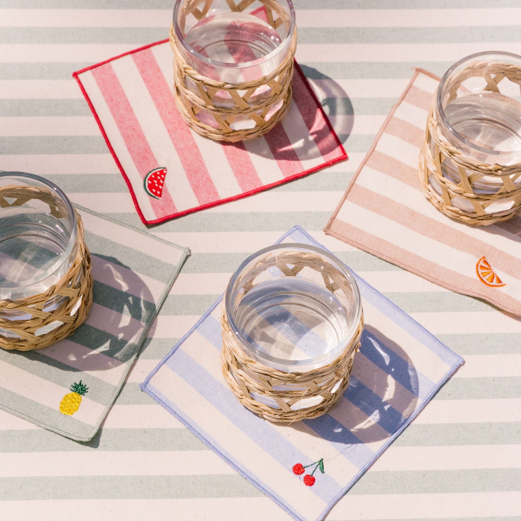 Wicker glasses on striped coasters with fruit designs on a table.