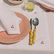 Dining table setting with pink plate, white napkin with cherry design, and cutlery on a white placemat.