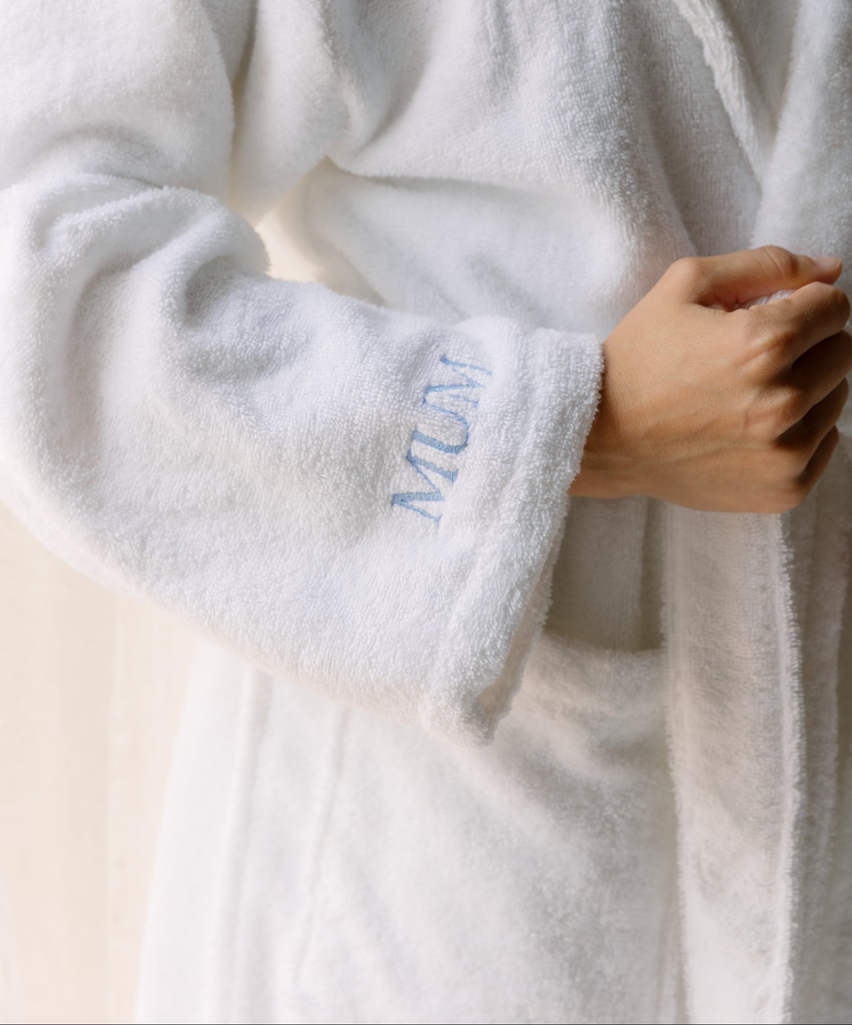 White robe with 'MUM' embroidery held by a hand against a neutral background